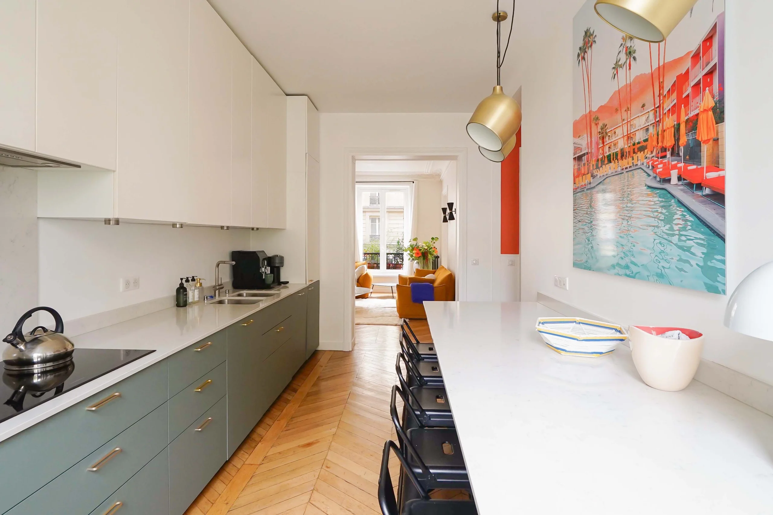 Cuisine haussmannienne avec meubles vert sauge, parquet chevron, luminaire doré et vue vers salon