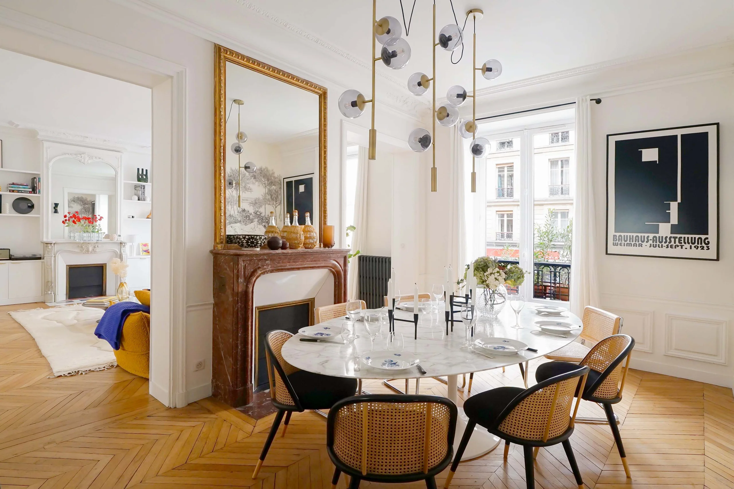 Salle à manger avec table ronde marbre blanc, chaises cannage bois clair, cheminée marbre, miroir doré, parquet chevrons et baies vitrées