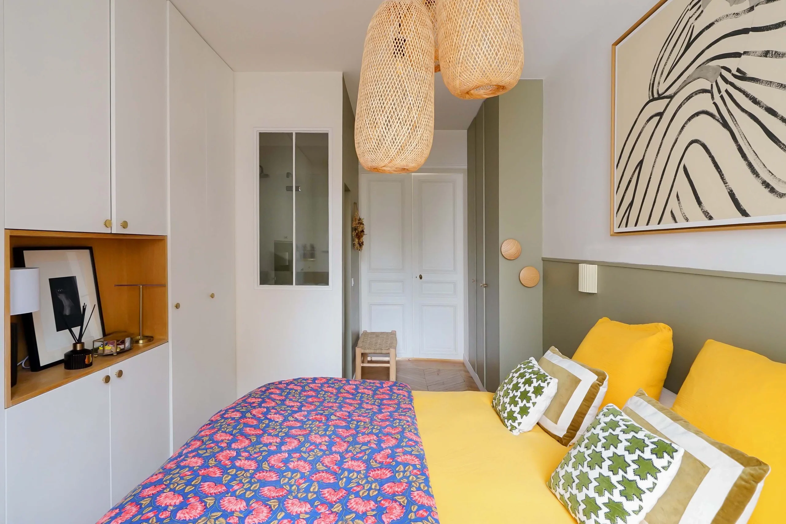 Chambre haussmannienne aux murs blancs, lit jaune avec tête de lit vert, suspension en rotin tissé, parquet et meuble suspendu en bois