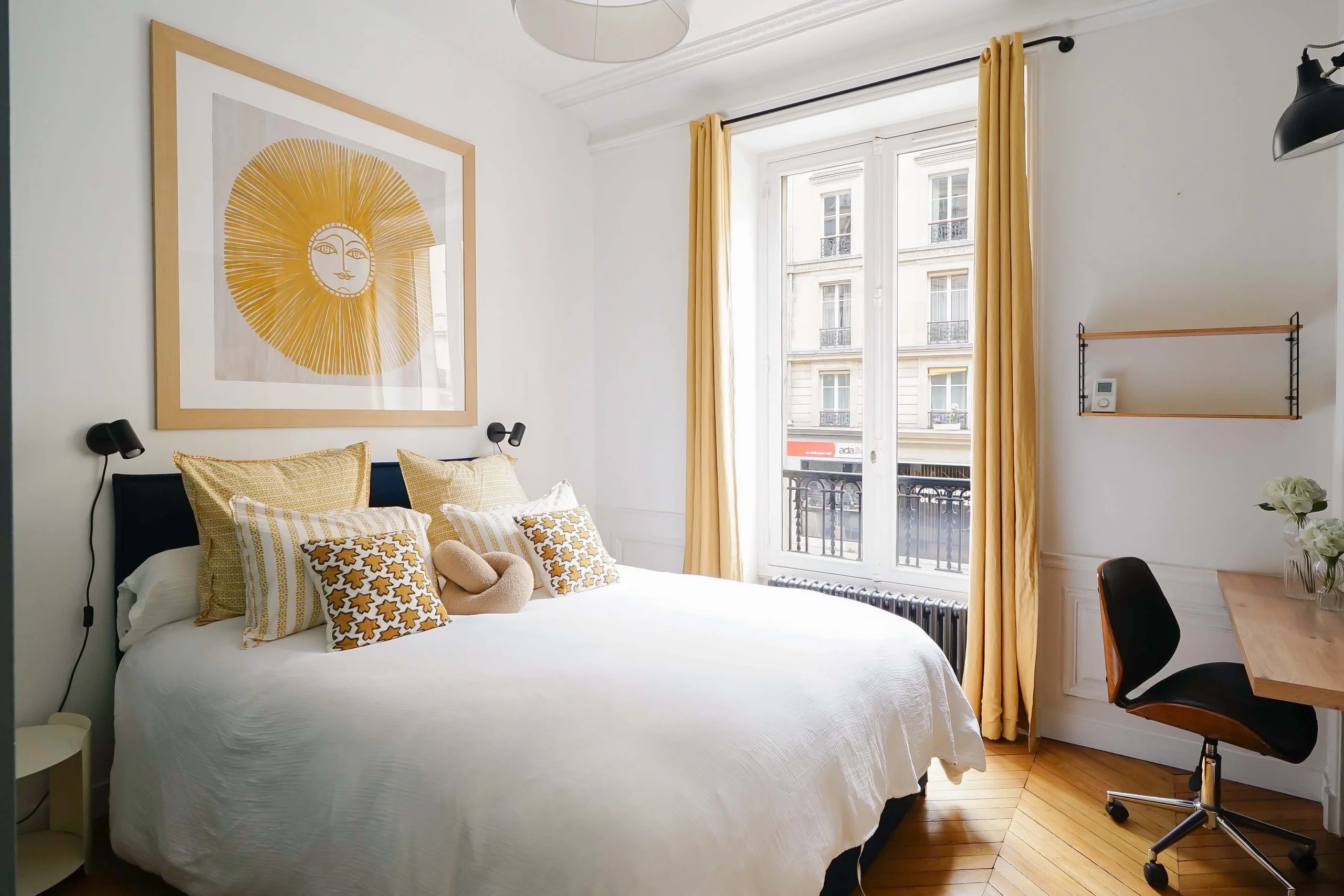 Chambre haussmannienne aux murs blancs, lit blanc avec coussins jaunes, rideaux dorés, parquet clair et fenêtre sur immeuble parisien