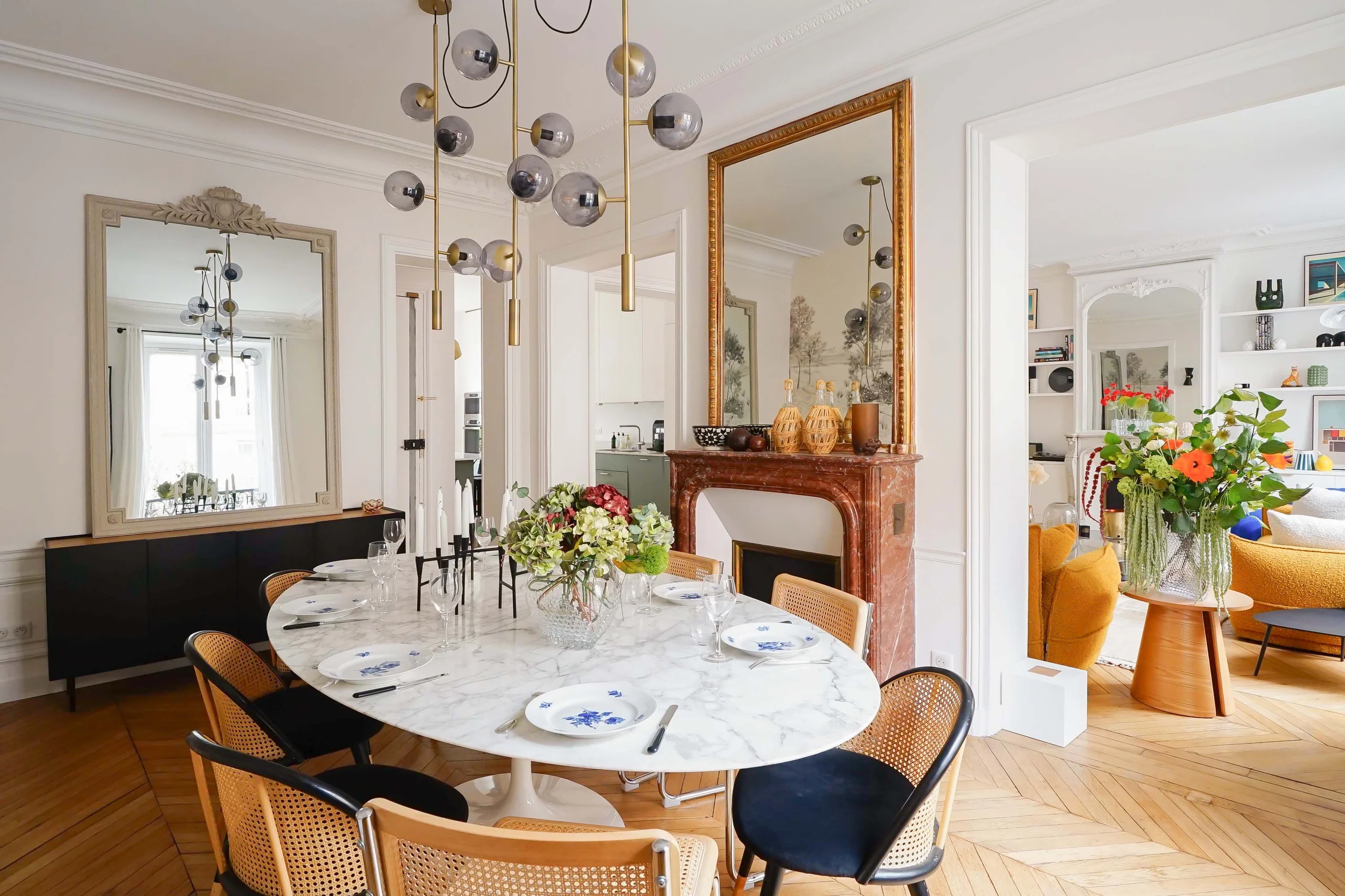 Salle à manger haussmannienne avec table ronde marbre, chaises cannage, cheminée bois, miroir doré, lustre design et parquet clair