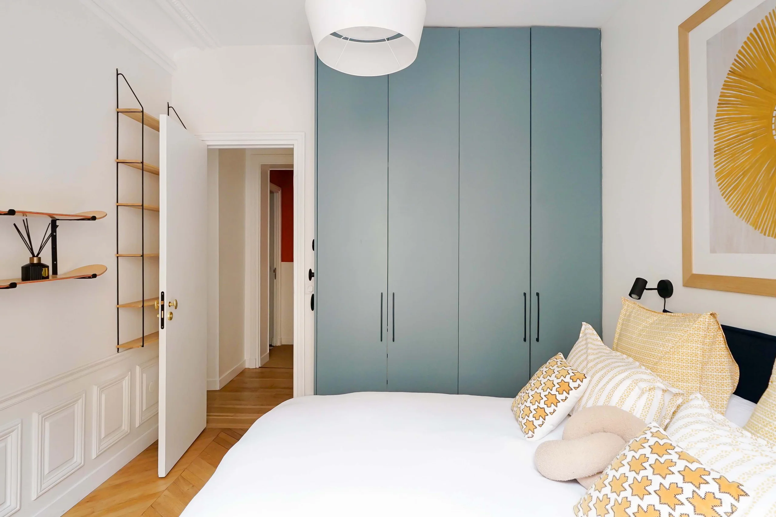 Chambre haussmannienne avec parquet bois, armoire bleue-gris trois portes, lit blanc et coussins colorés, luminaire plafond