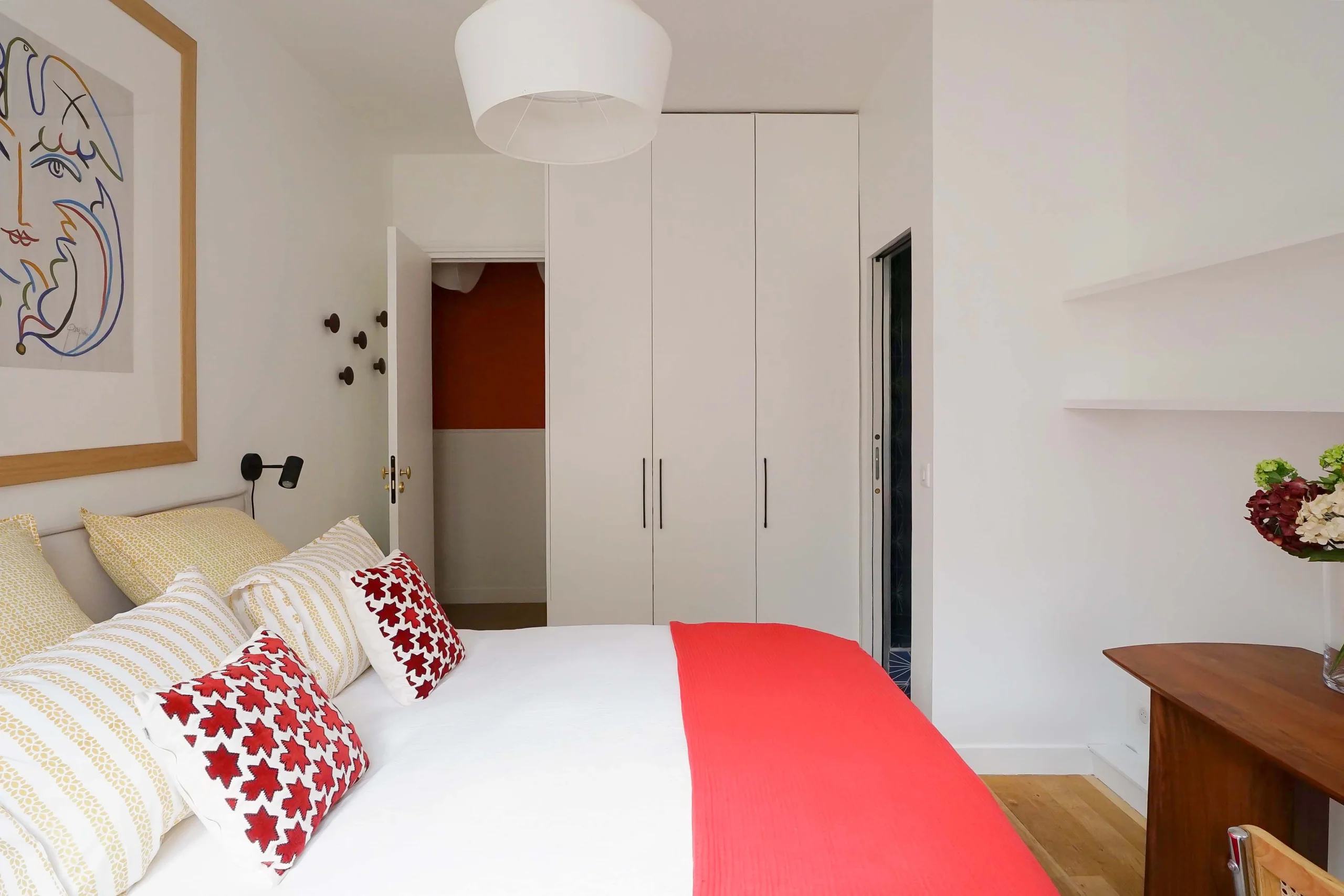 Chambre ensoleillée avec lit blanc, coussins rouges, placards blancs et plafond haut haussmannien