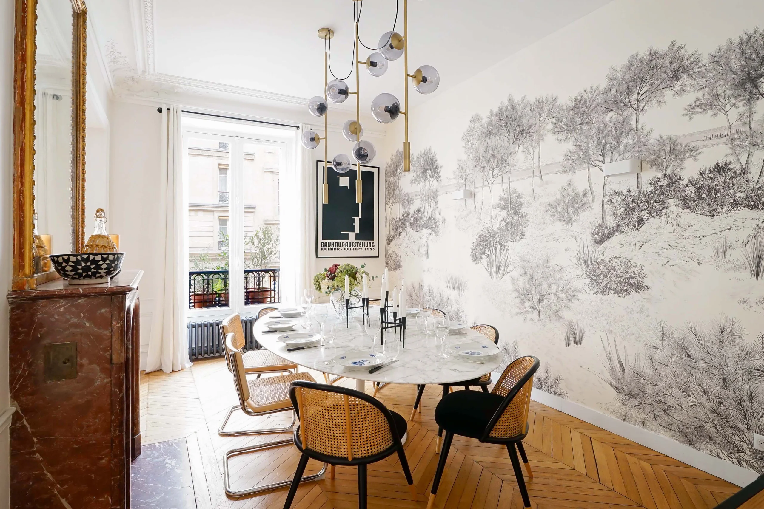 Salle à manger haussmannienne avec table marbre, chaises cannées, papier peint forêt grise et parquet chevron