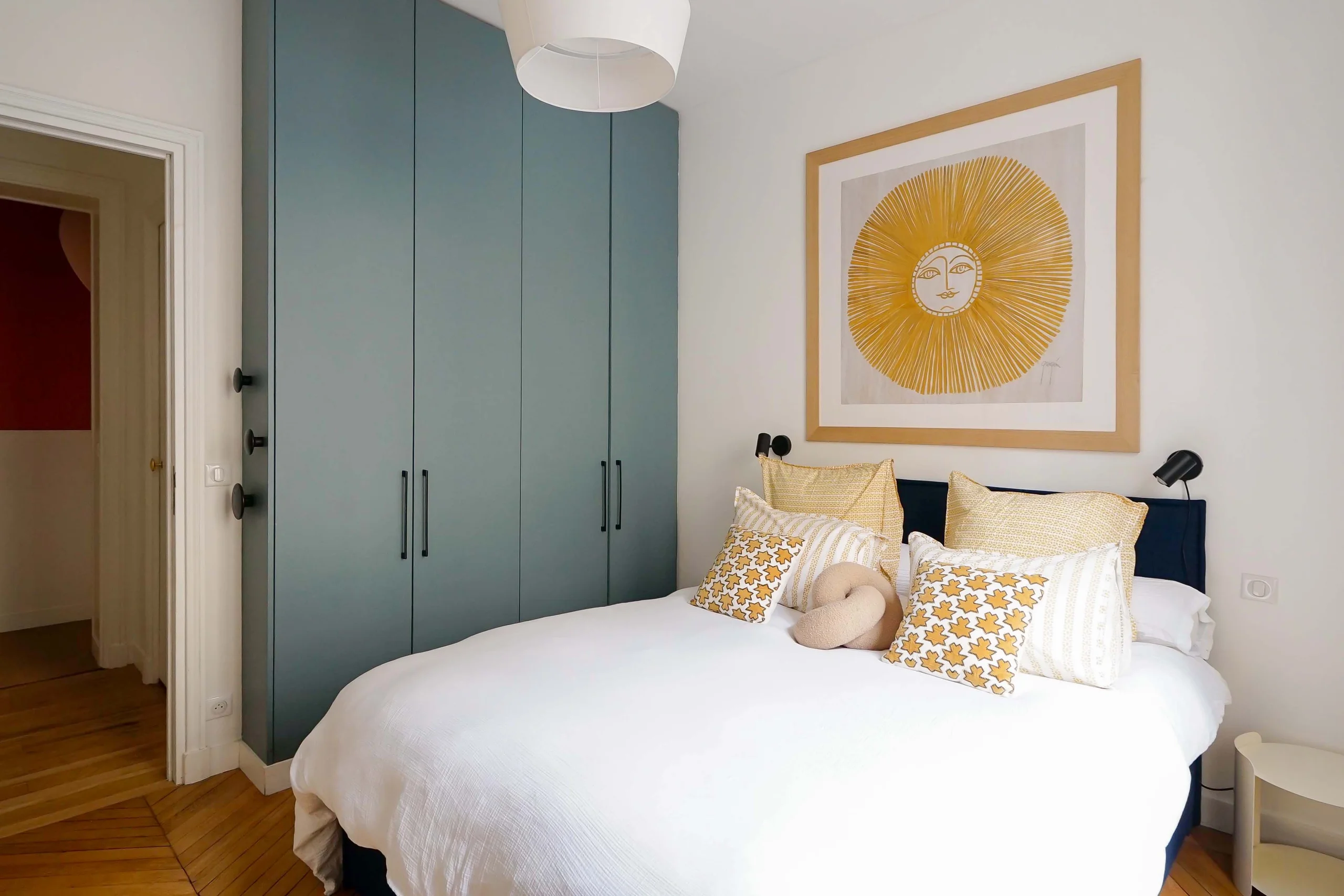 Chambre avec lit blanc, armoire bleu-gris et parquet bois clair, œuvre jaune au mur