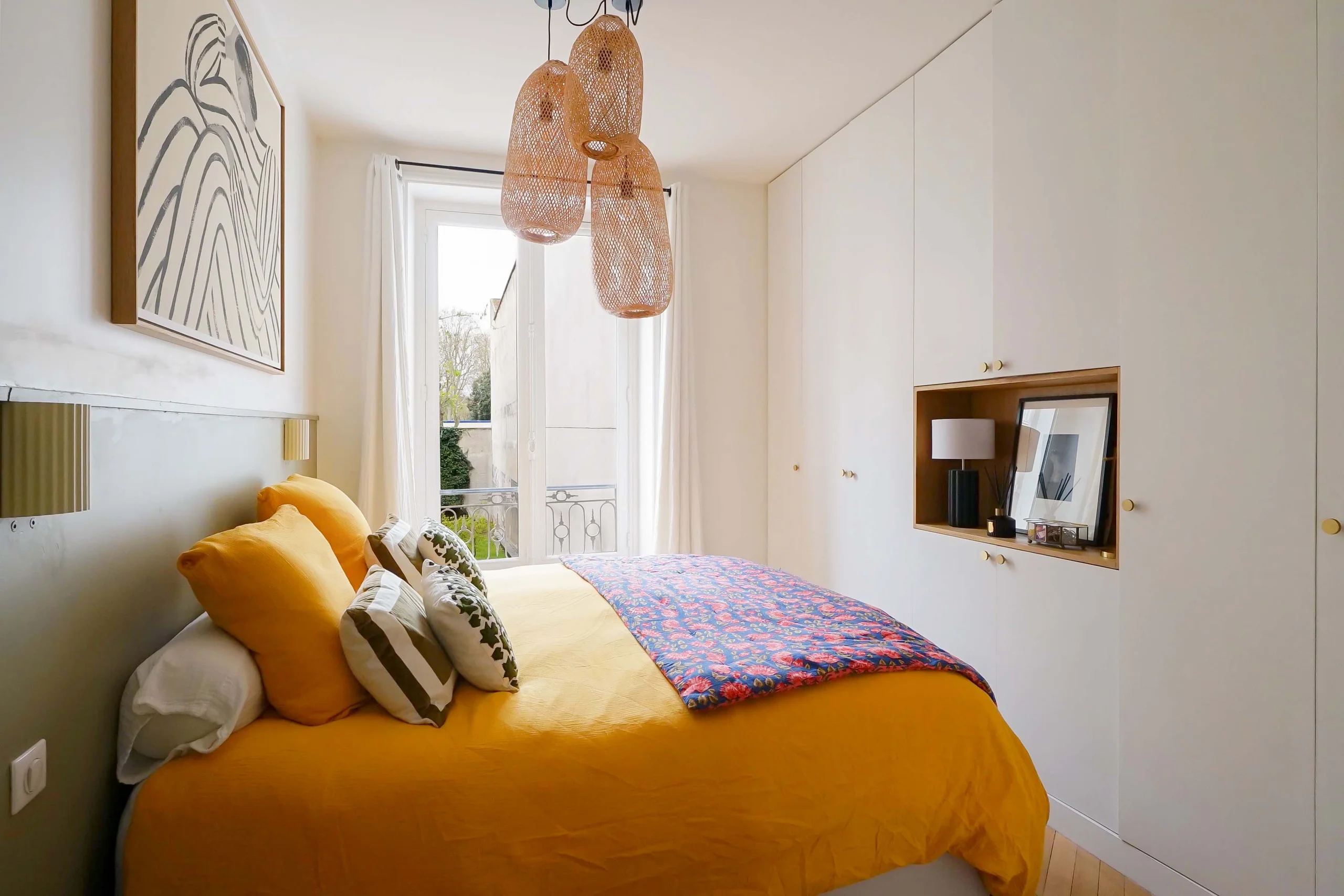 Chambre haussmannienne avec lit ocre, coussins jaunes, suspension rotin, portes-fenêtres et parquet clair