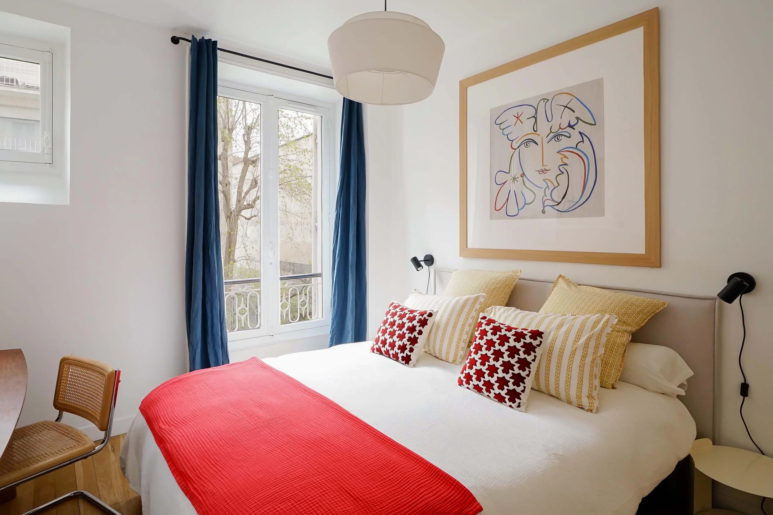 Chambre haussmannienne lumineuse avec lit blanc, plaid rouge, coussins colorés, fenêtres hautes et rideaux bleus