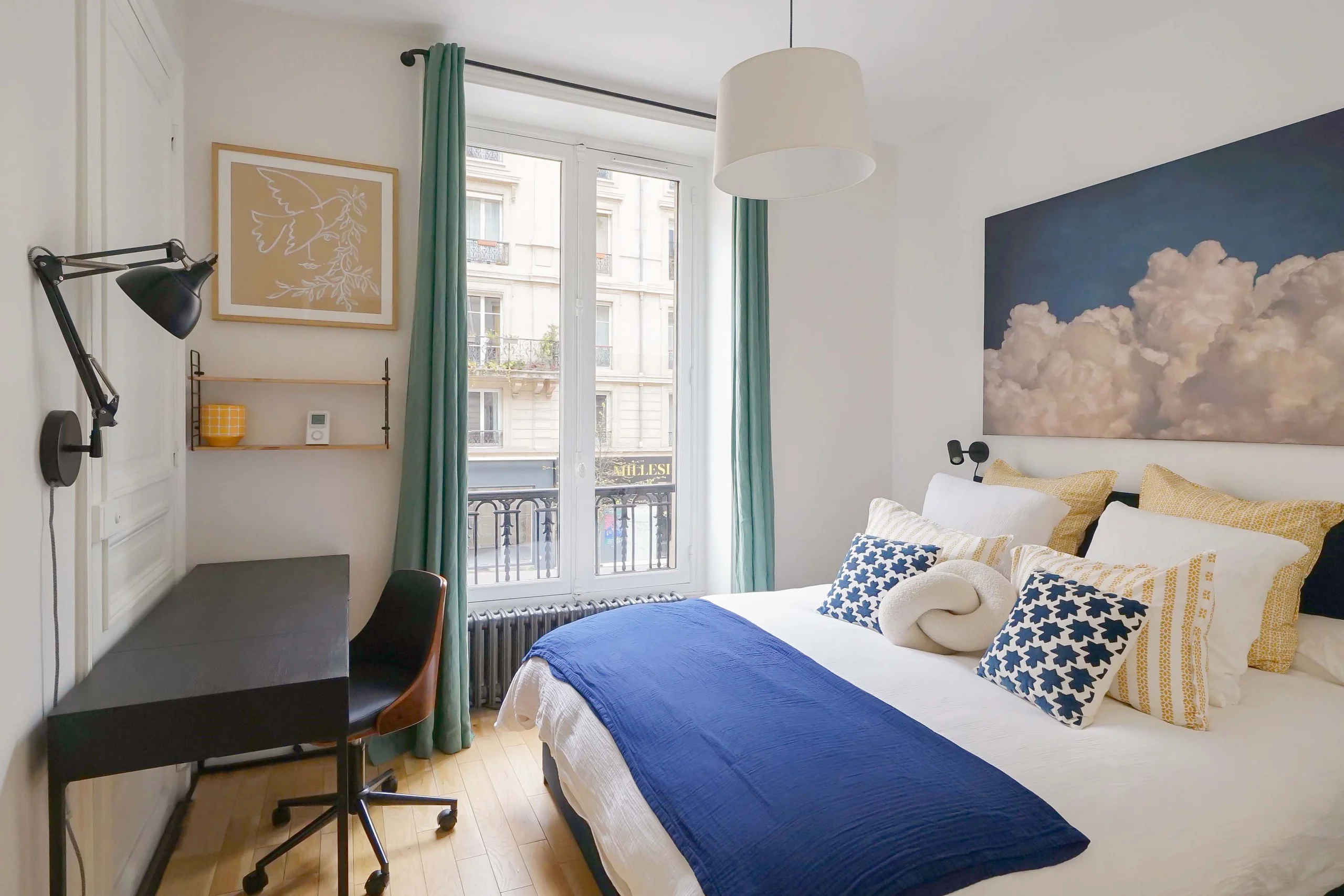 Chambre haussmannienne avec lit blanc, jeté bleu, rideaux verts, bureau noir et fenêtres donnant sur Jardin du Luxembourg