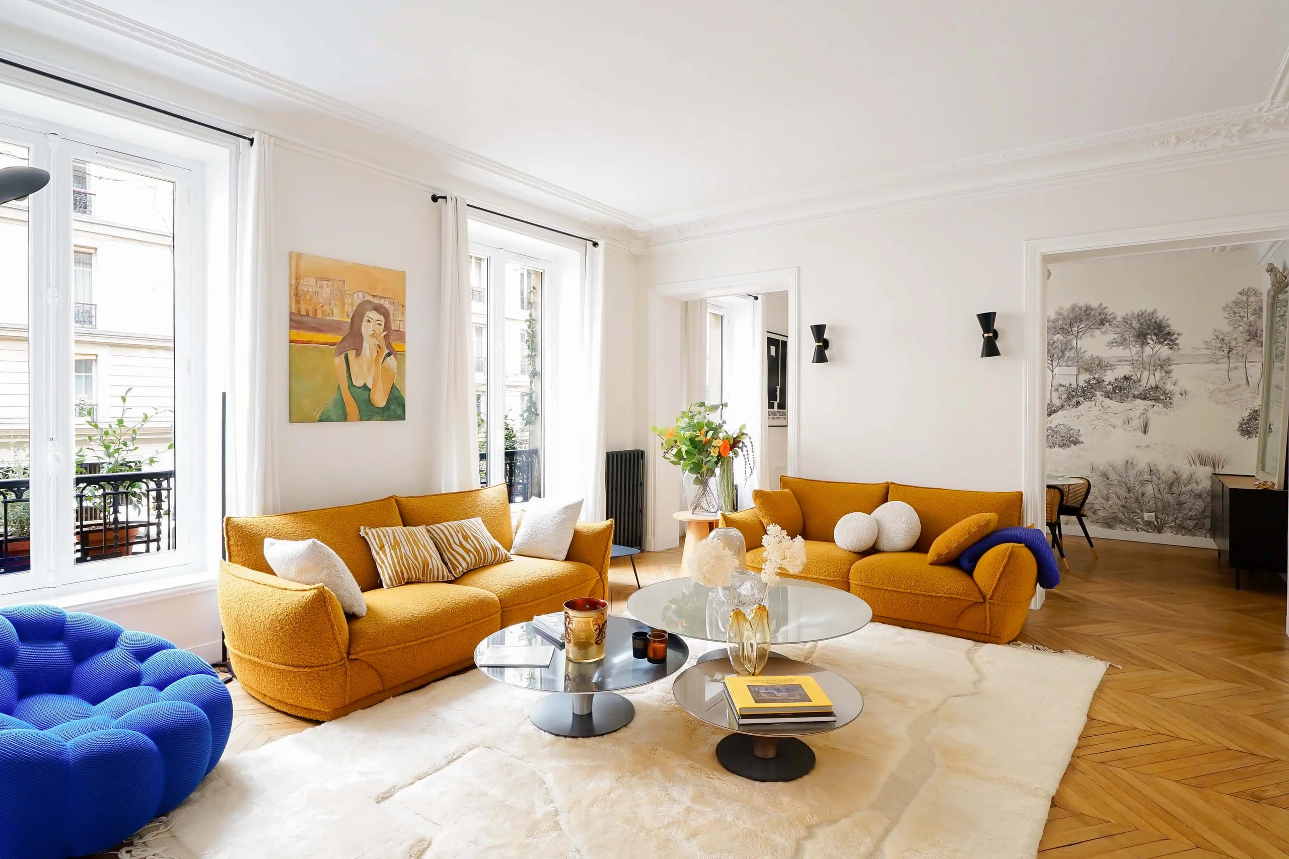 Salon haussmannien lumineux avec canapés jaune ocre, parquet clair, grandes fenêtres et pouf bleu électrique