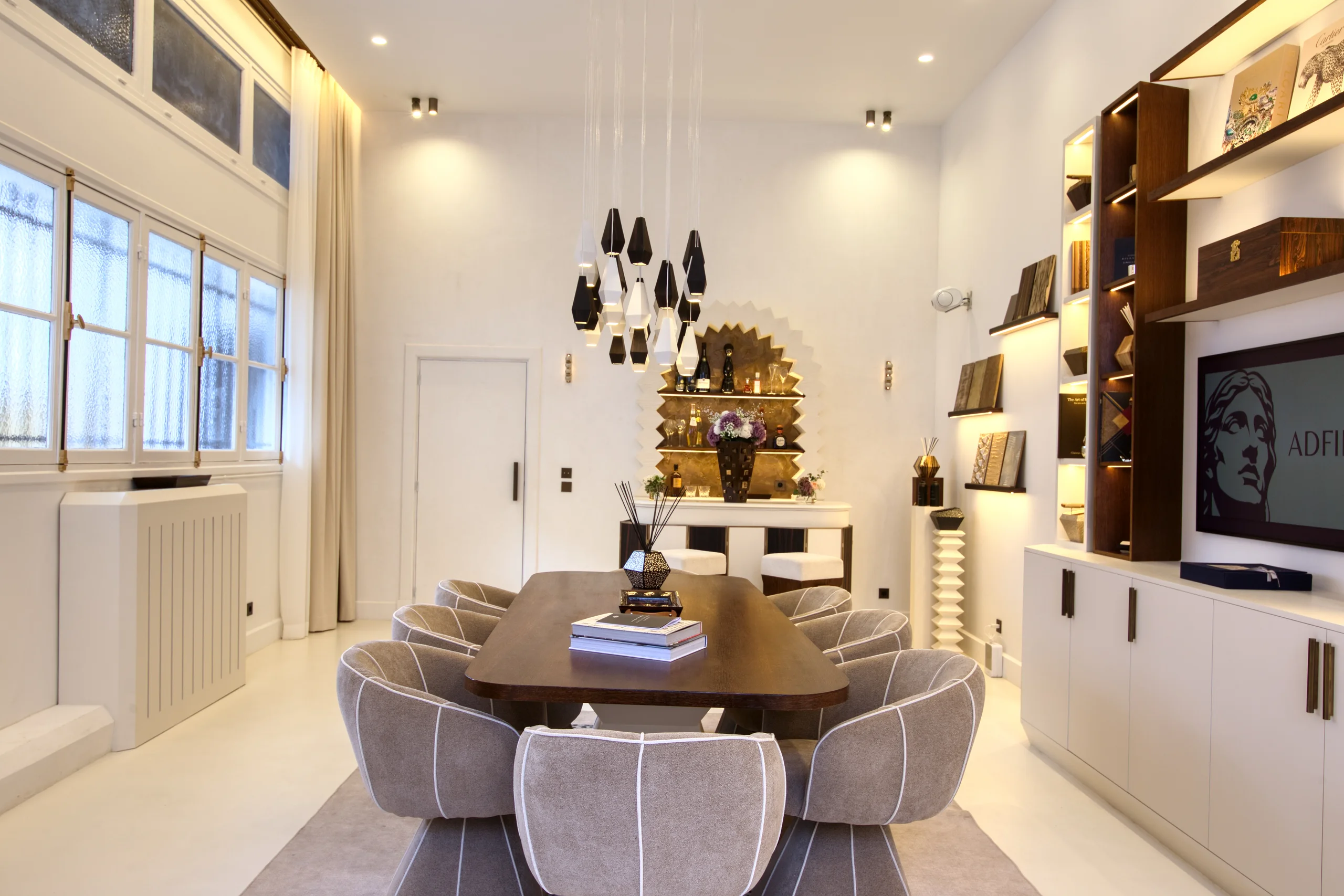 Grand salon avec table ovale bois massif, chaises grises, étagères bois intégrées, lustre suspendu noir, lumière naturelle des fenêtres
