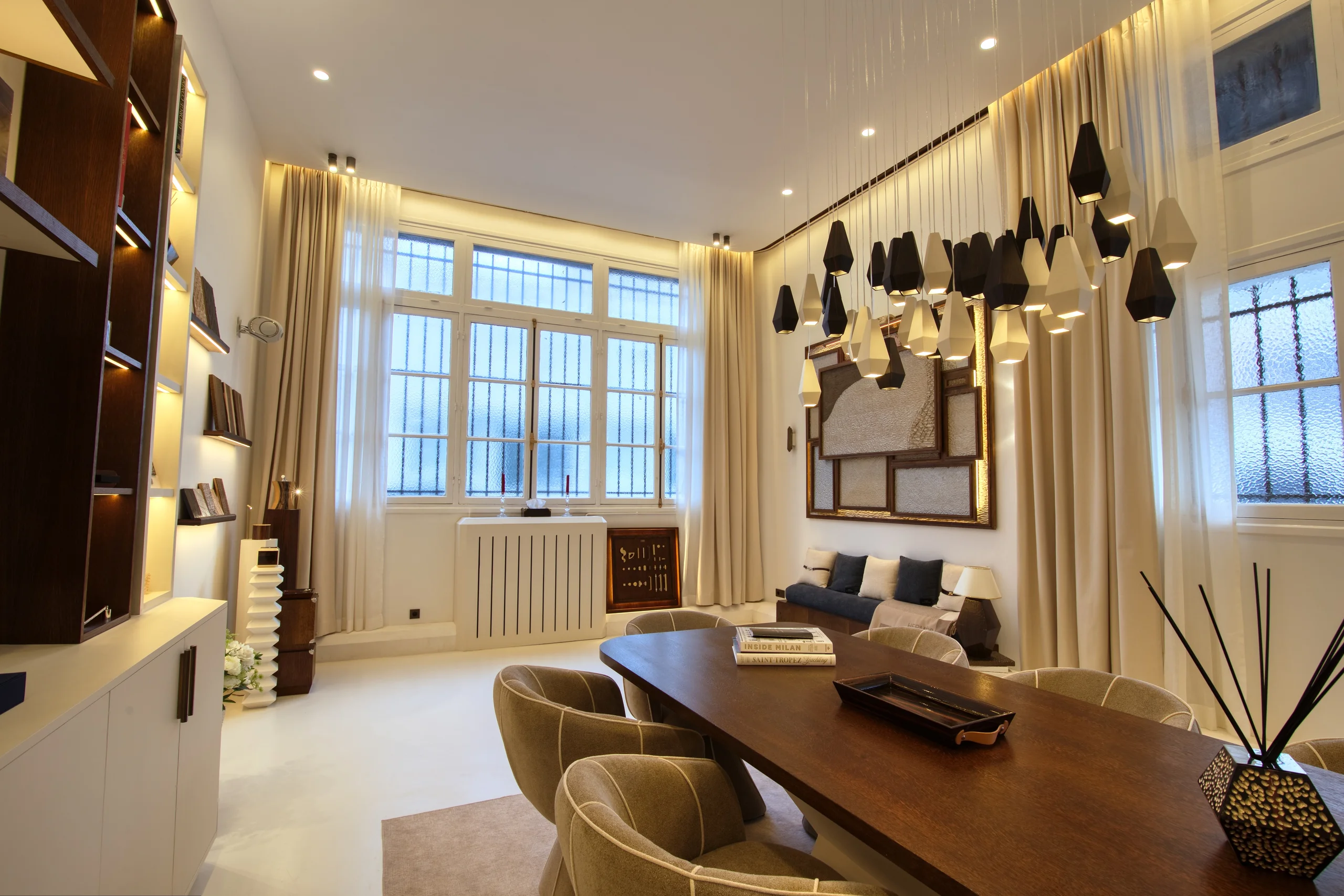 Grand Salon avec luminaires suspendus noirs, grandes fenêtres, rideaux crème, mobilier design et éclairage ambiant chaleureux