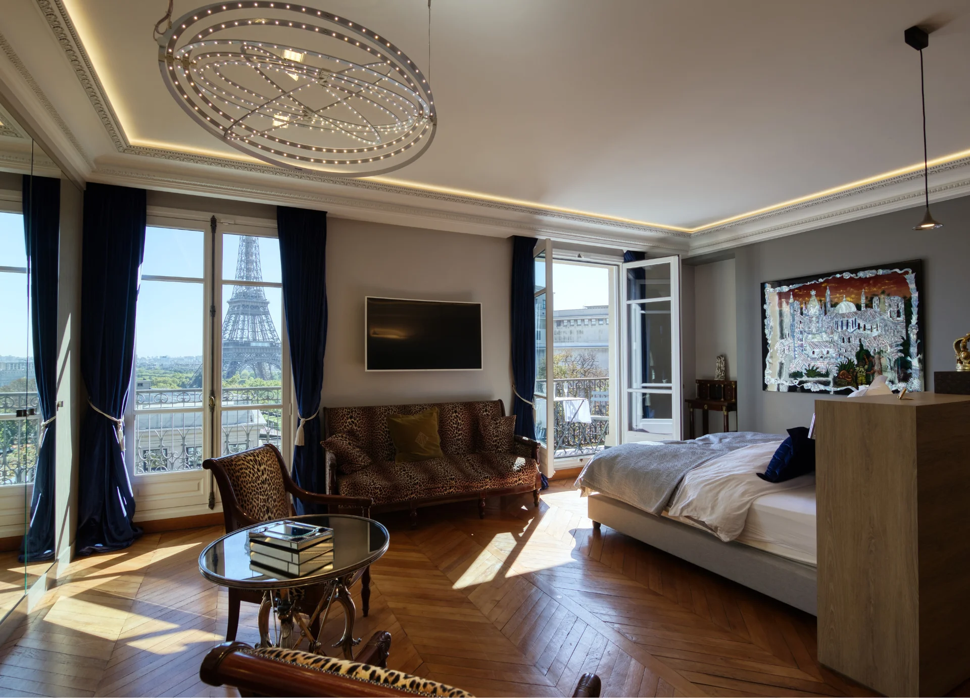 Suite spacieuse avec parquet bois, canapé cuir cognac, vue panoramique Tour Eiffel par baies vitrées, plafond blanc avec lustre géométrique