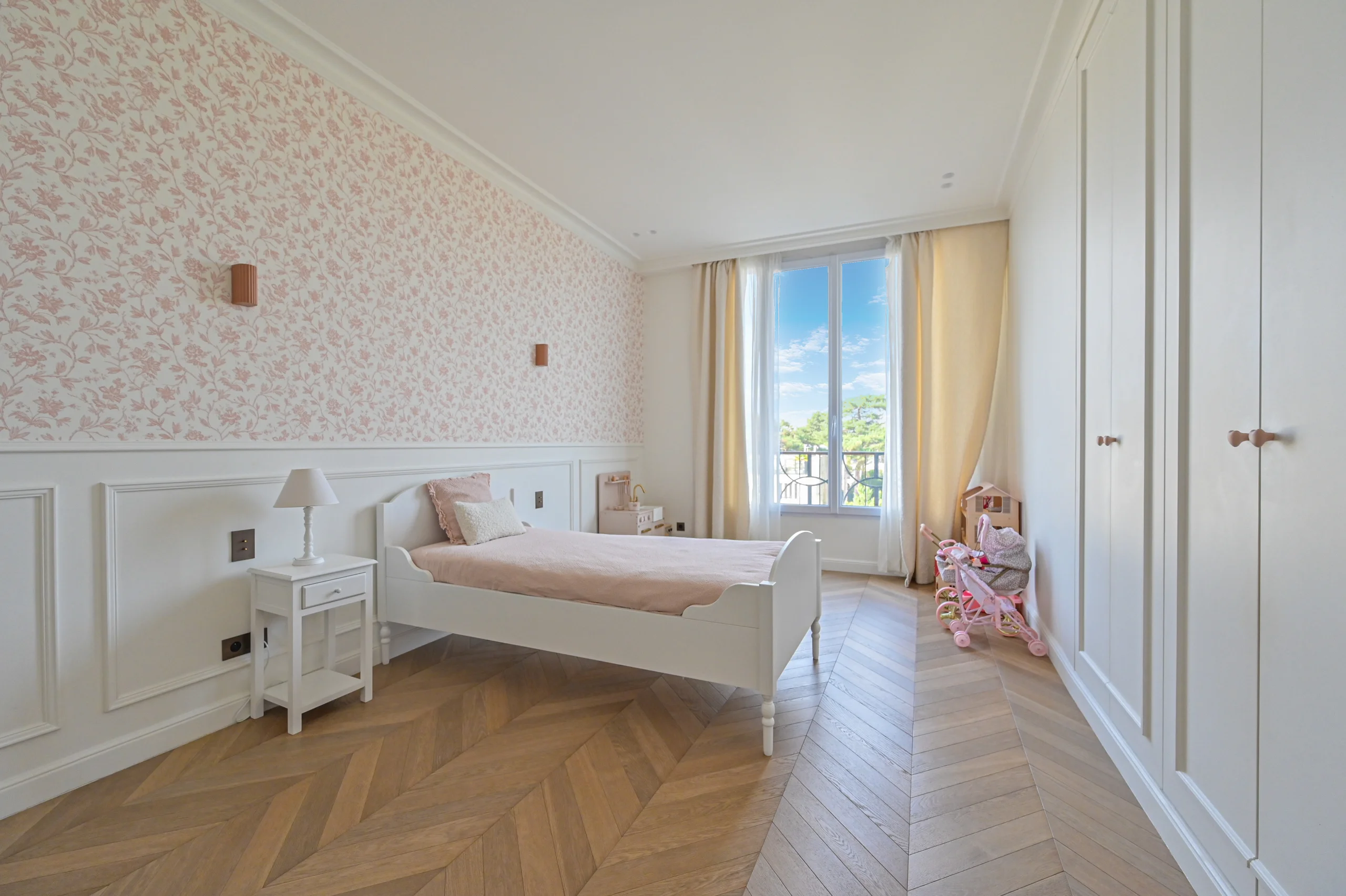 Chambre lumineuse avec parquet chevron, lit blanc, murs rose poudré et accès terrasse ensoleillée