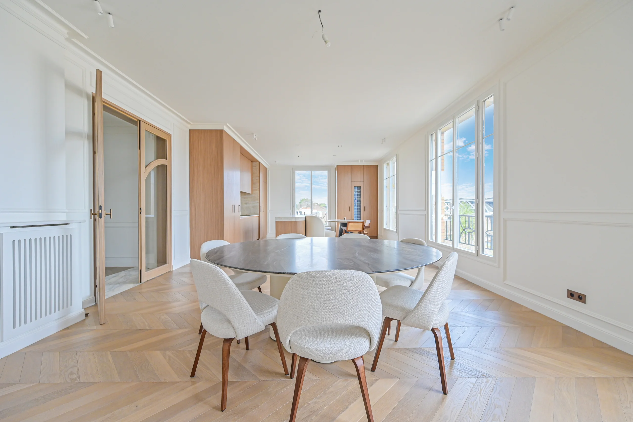 Salle à manger lumineuse avec table ronde blanche, chaises grises, parquet clair et baies vitrées donnant sur Roland-Garros