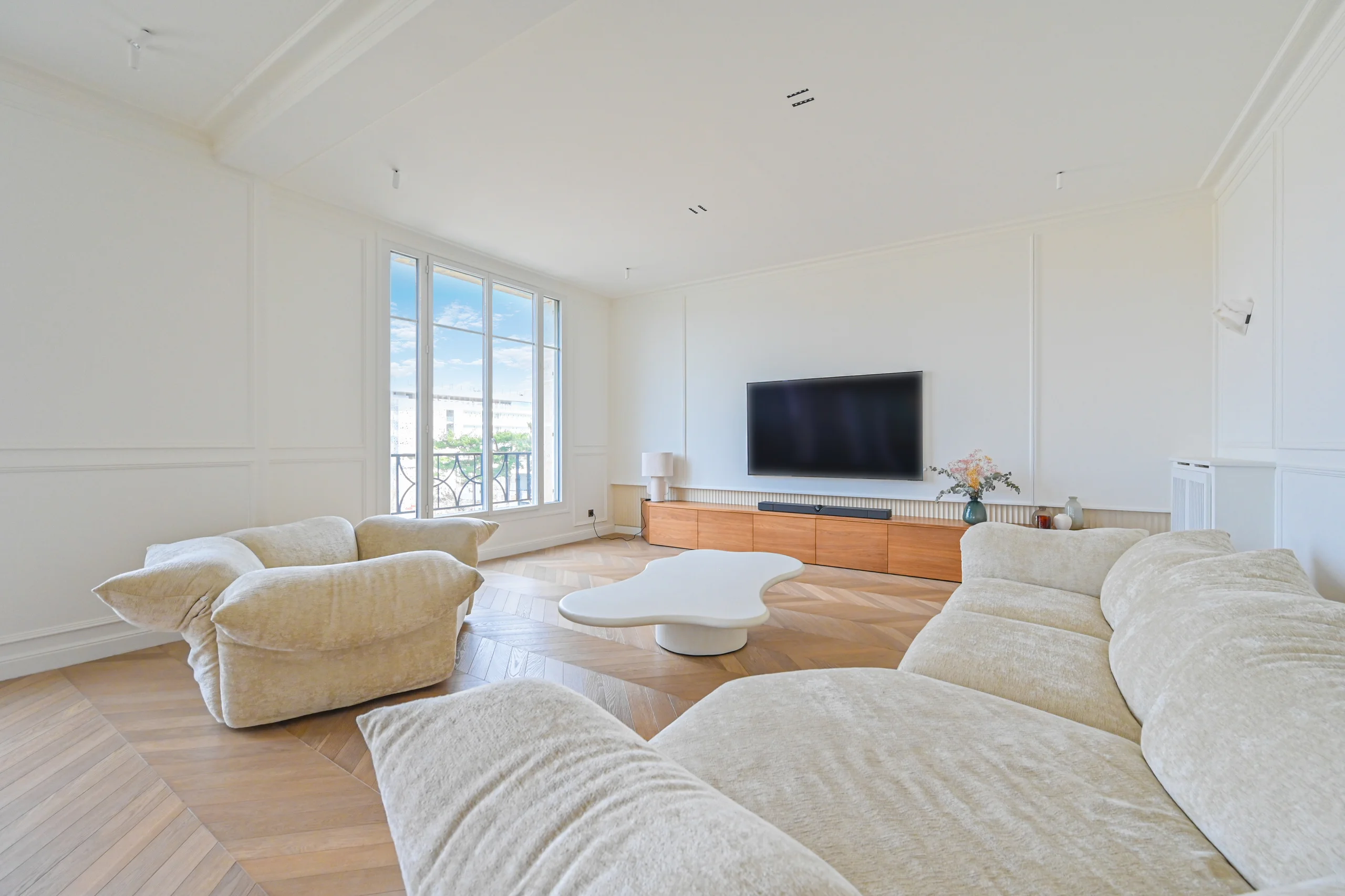 Salon lumineux aux murs blancs, canapés beige, sol parquet, télévision sur meuble bois clair, baie vitrée