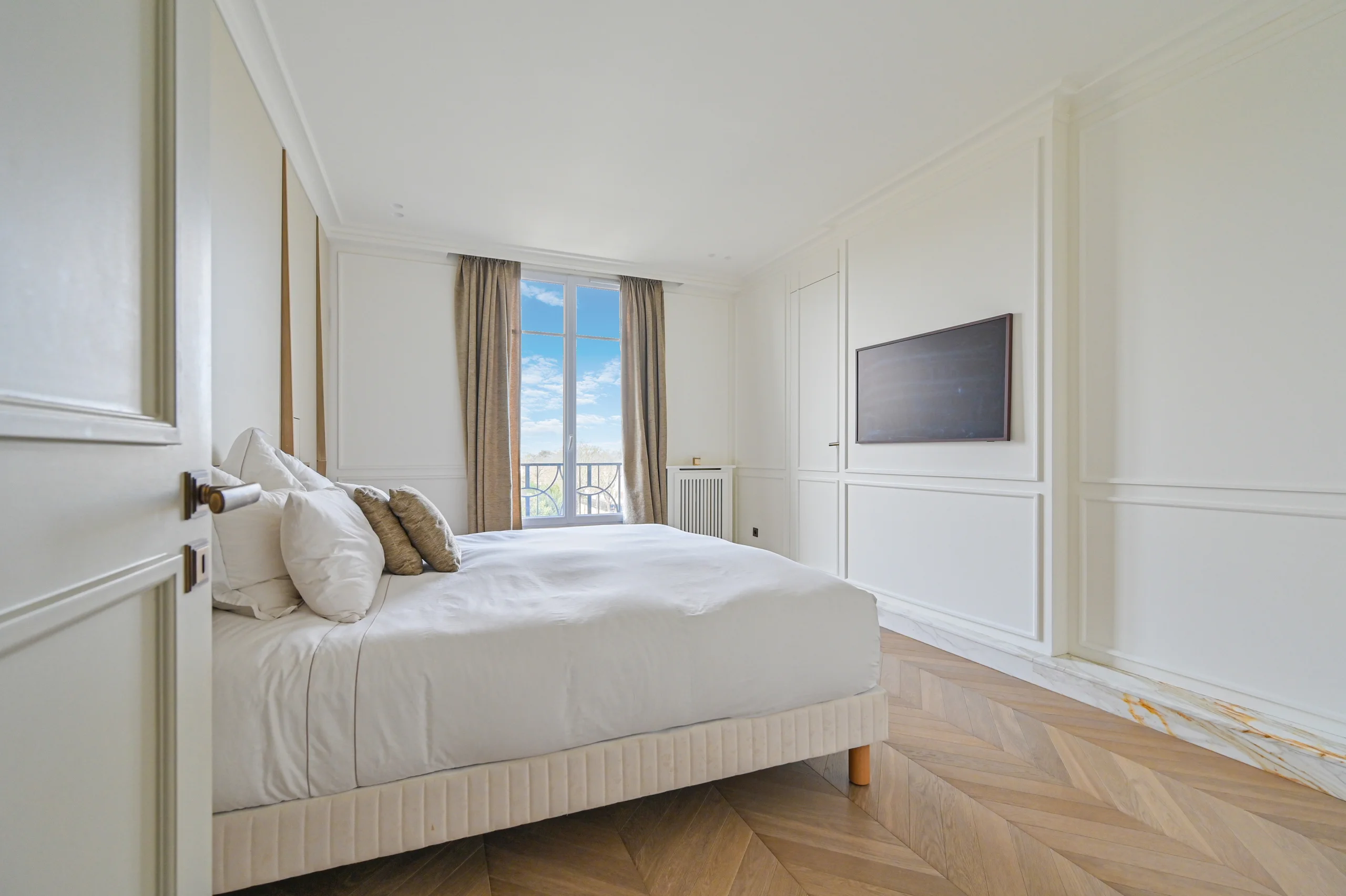 Chambre blanche lumineuse avec lit crème, grandes fenêtres, parquet clair et écran mural
