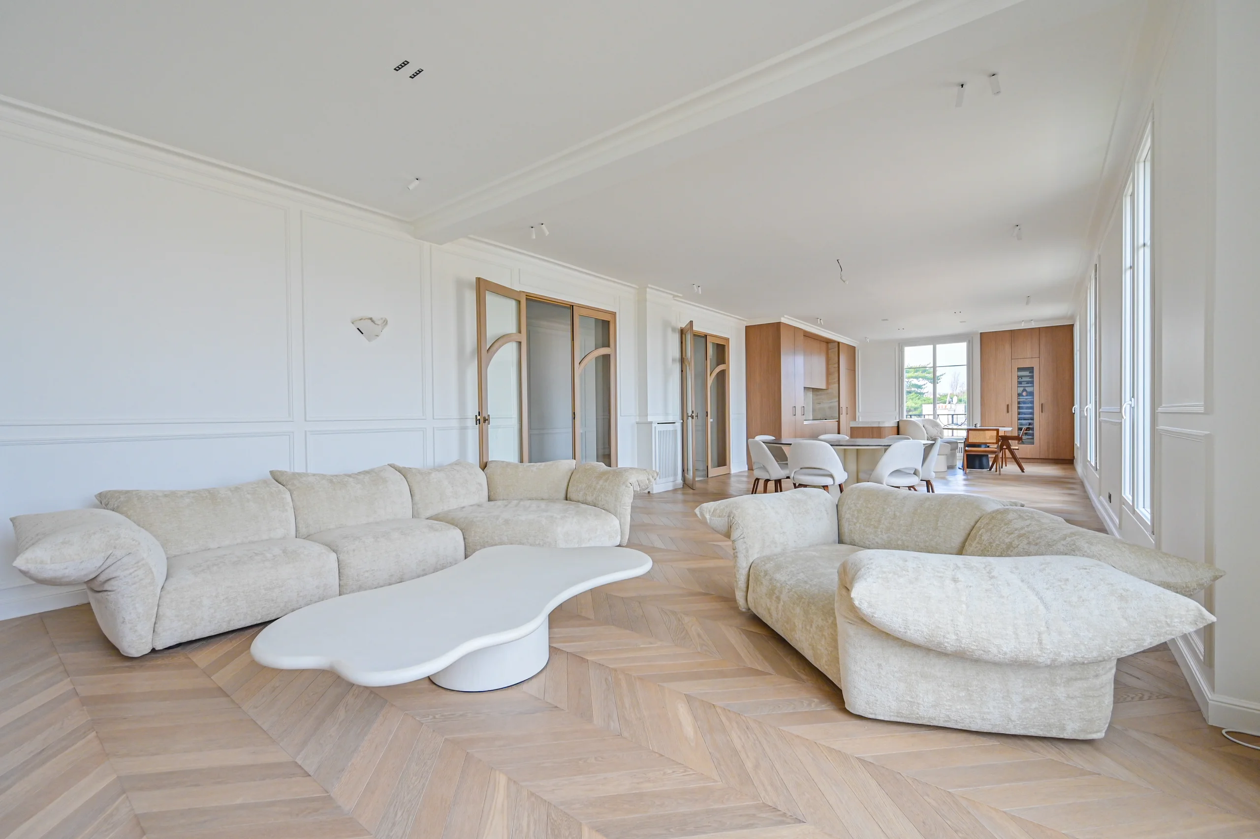 Salon spacieux aux murs blancs, parquet chevron clair, canapés arrondis blanc crème et table basse blanche minimaliste