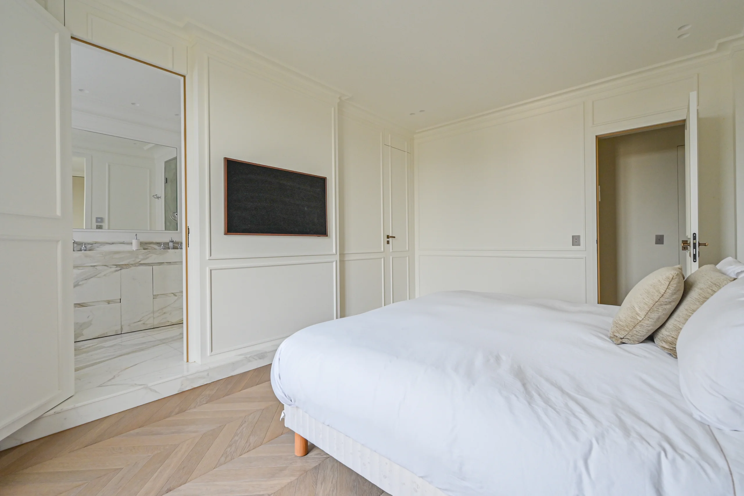 Chambre épurée aux murs blancs, lit blanc, parquet clair, écran plat anthracite et salle de bains ouverte
