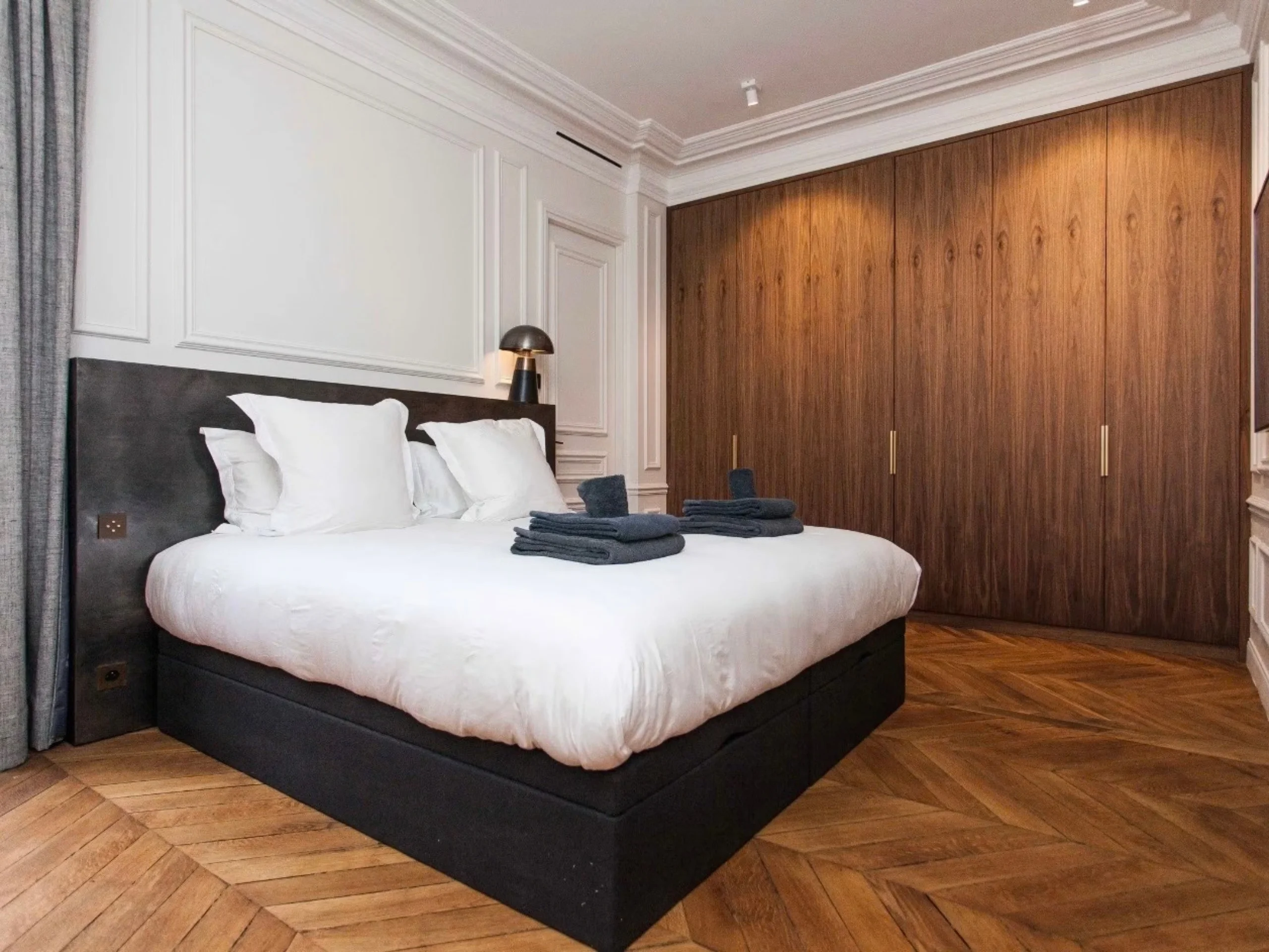 Chambre avec lit noir aux draps blancs, placards bois foncé, parquet chevron et plafond moulures