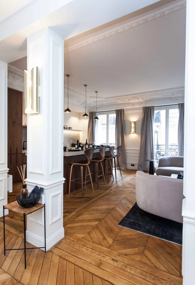 Séjour haussmannien aux murs blancs, parquet point de Hongrie, colonnes blanches, cuisine noire ouverte et lustre pendant