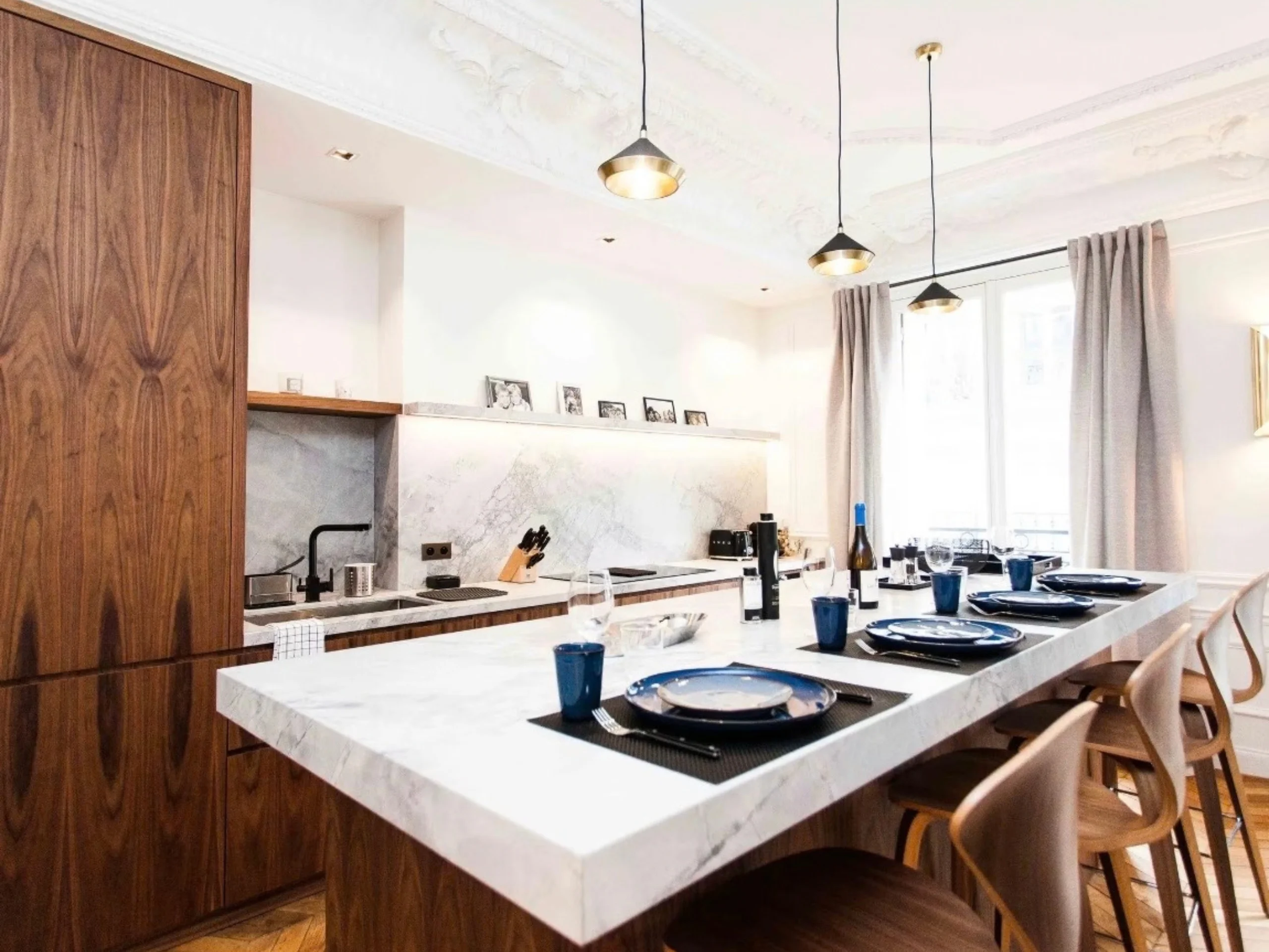 Cuisine ouverte avec îlot marbre blanc, placards bois massif, luminaires dorés suspendus et salle à manger attenante