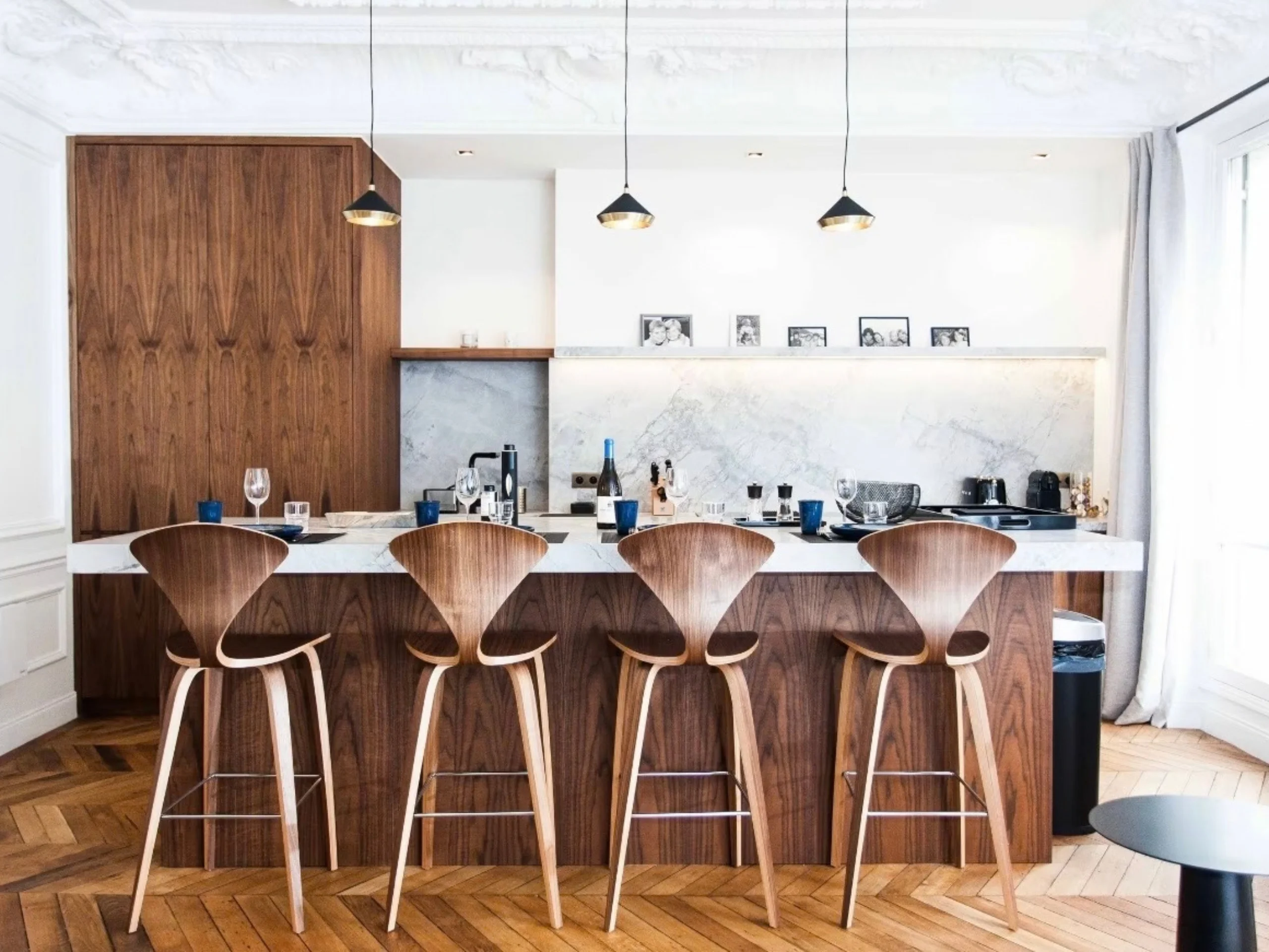 Cuisine ouverte avec îlot en bois massif, quatre tabourets design, carrelage marbre blanc et suspensions dorées
