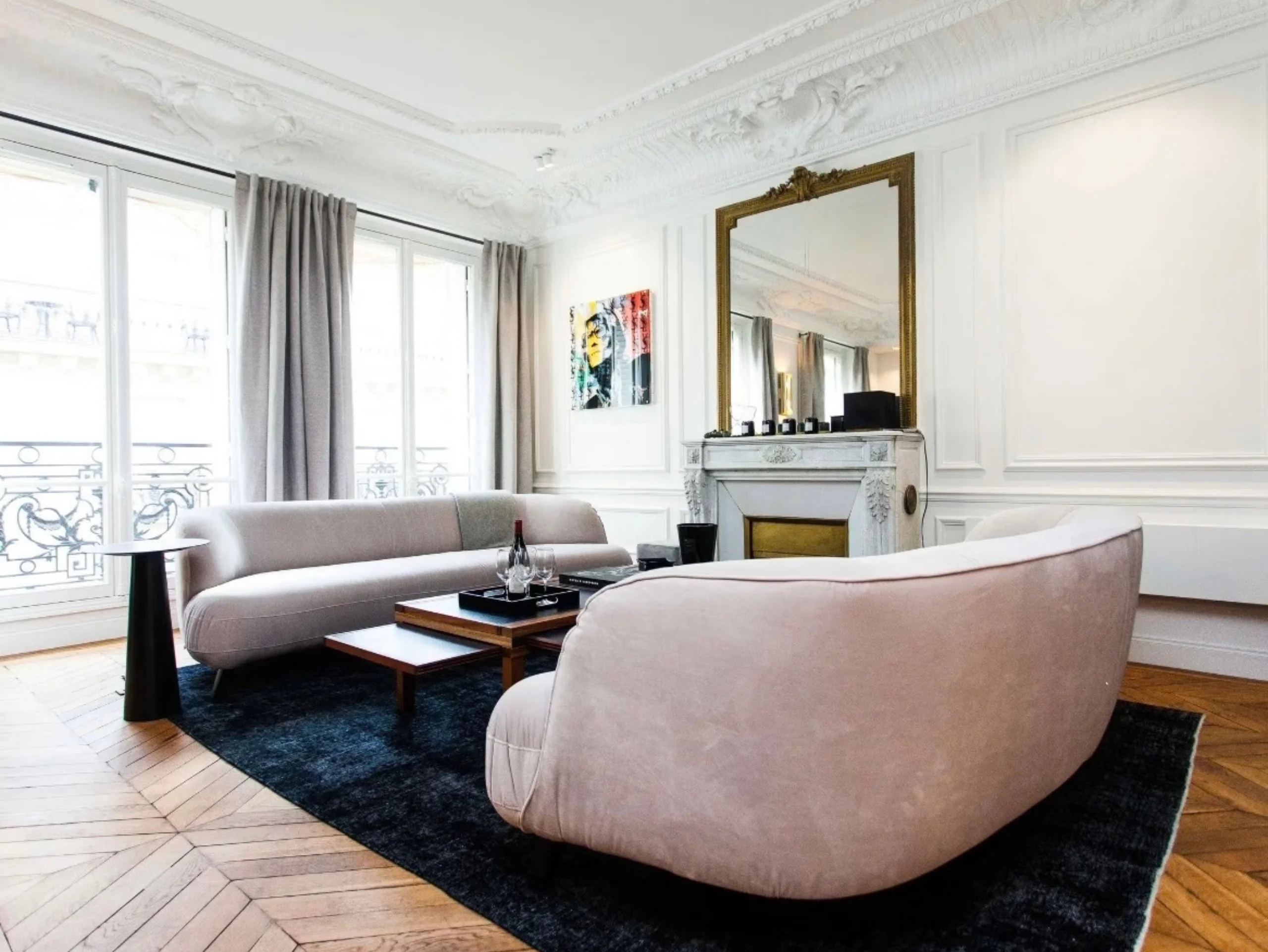 Salon haussmannien lumineux avec canapés arrondis rose poudré, parquet chevron, miroir doré et hautes fenêtres classiques